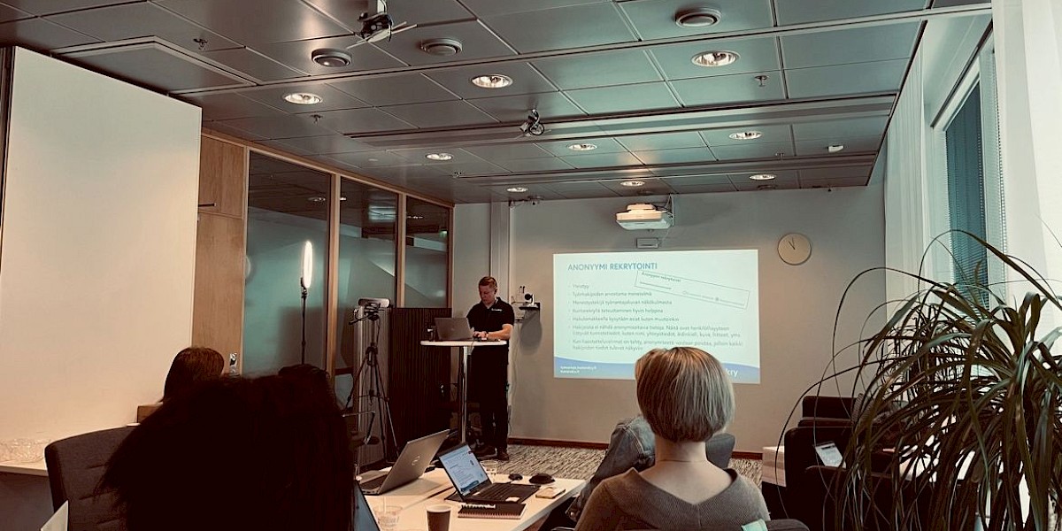 Valokuva kokoustilasta, jossa yksi henkilö seisoo huoneen takaosassa edessään pöytä, jossa tietokone, josta heijastuu esitys takaseinälle. Etualalla oikealla viherkasvi, keskellä henkilö selkä kohti kameraa.